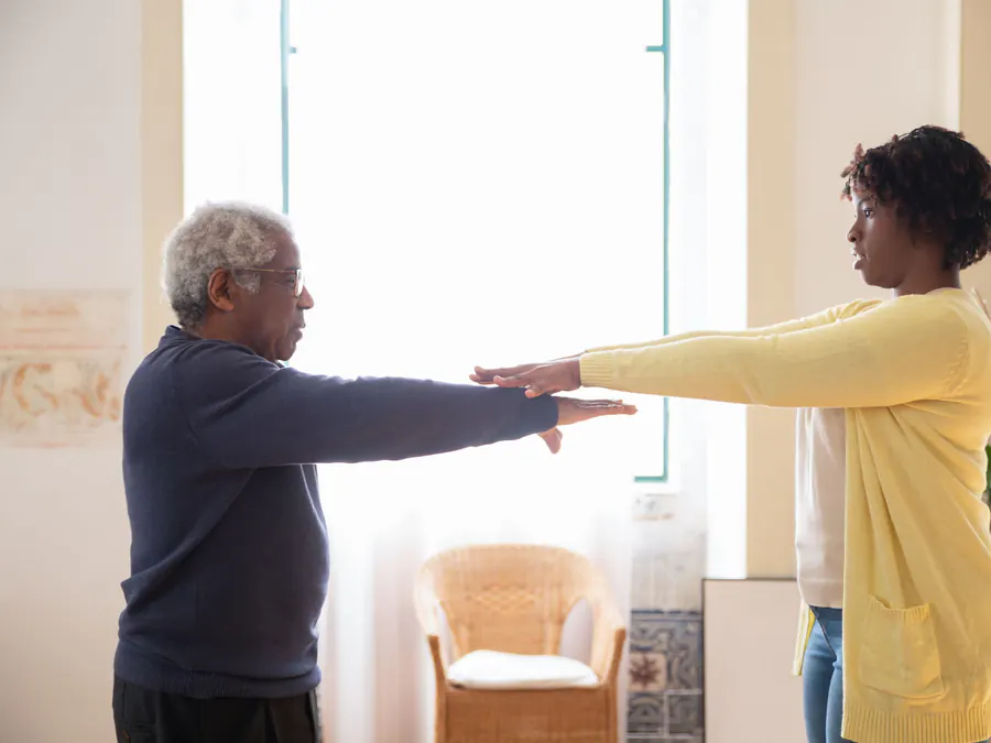 Older man doing guided arm-raise exercises with a therapist in his sunny living room