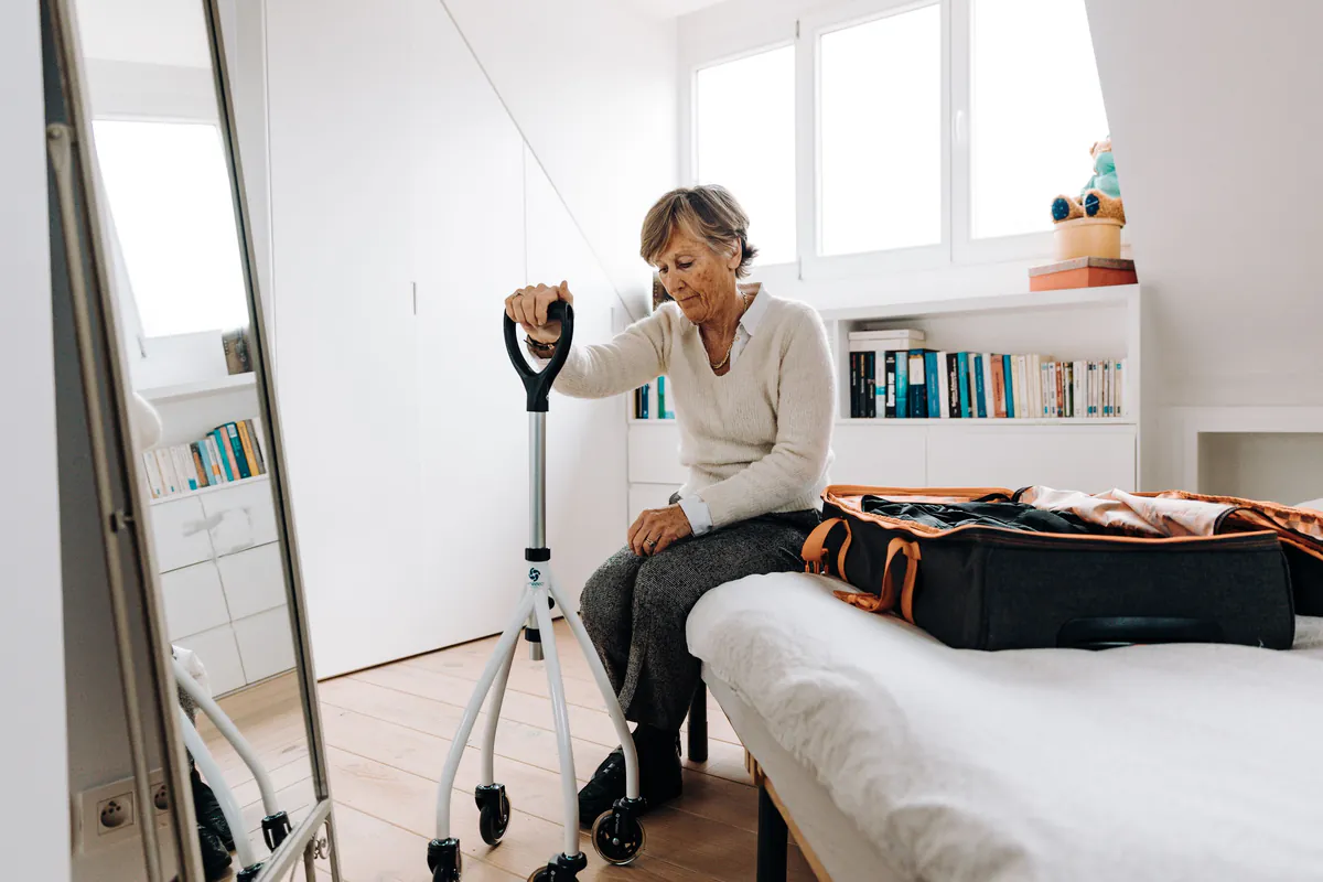 Older woman sitting on her bed beside a four-wheeled rollator walking aid in a bright bedroom