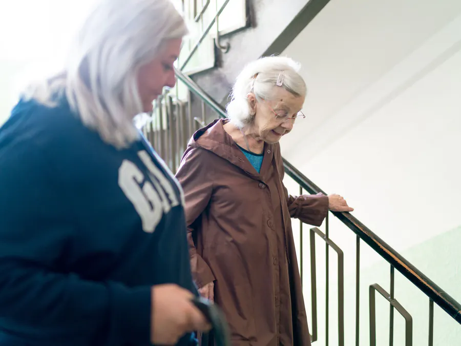 Carer steadying an older woman as she walks down the stairs at home, holding the handrail